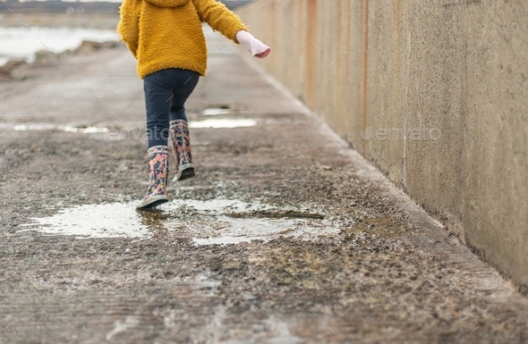A little girl with rubber boots runs into a puddle on a rainy day