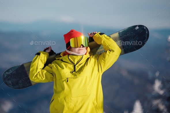 Snowboard up raise. Yellow, mustard jacket, red knitted hat, ski goggles.