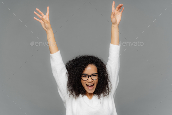 woman raises hands up, being in high spirit, dances over grey background