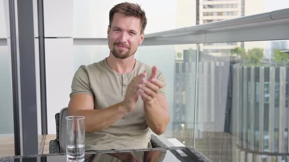 A Young Man Applauds the Camera and Nods with a Smile As He Sits on a Balcony in a Apartment alt