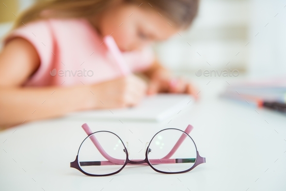 Little girl is writes in school notebook, blurry image. Glasses for vision in focus. Poor vision
