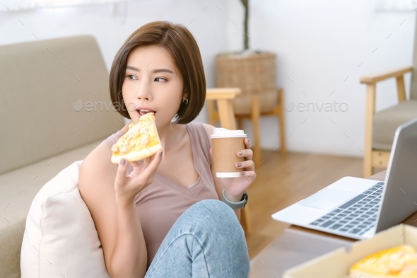Smiling beautiful Asian woman having lunch while working on laptop computer in living room at home.