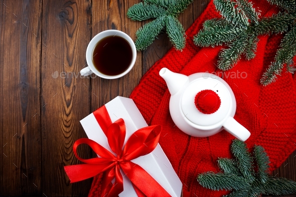 On a wooden background there is a red scarf and there is a teapot and a cup of tea
