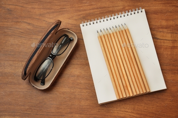The top view flat lay of workspace. Eyeglasses in eyeglasses case and notepad with pencils.