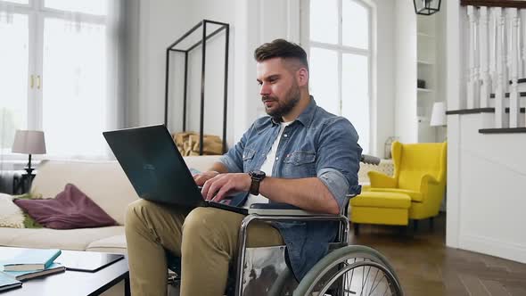 Modern Man with Beard Sitting in Wheelchair and Remotely Working at Home on Laptop alt