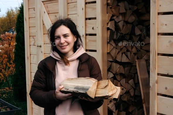 Happy young caucasian woman holding firewood at the.shed in the backyard