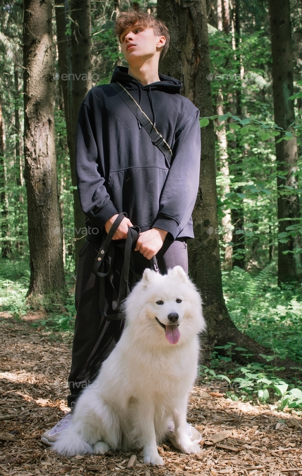 A young man stands with a white fluffy Samoyed dog on a leash in a summer forest and looks away