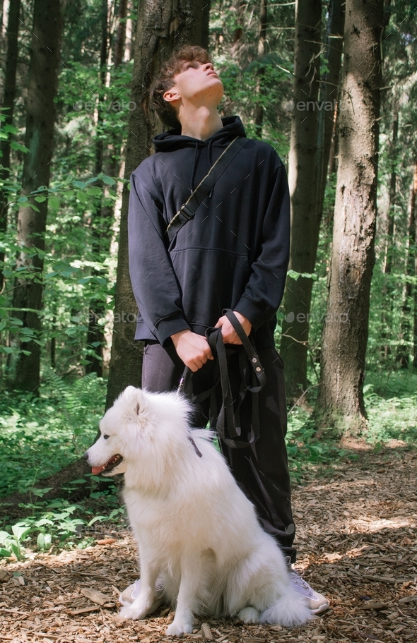 A young man stands with a white fluffy Samoyed dog in a summer forest