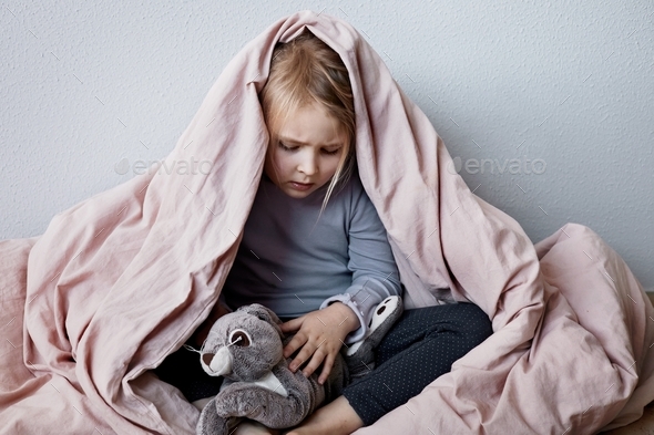 Little scared girl hiding under blanket with her favorite toy. Home violence, fear, war, psychology