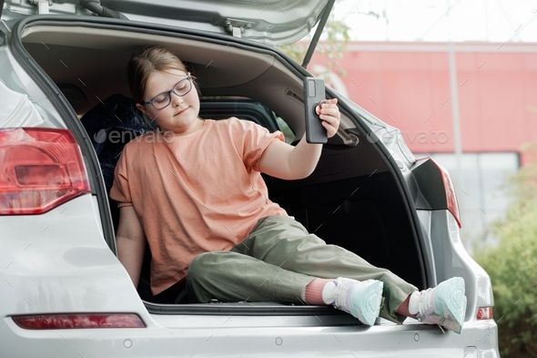 Nothing else matters: preteen girl sitting in car trunk making selfie ignoring her surroundings