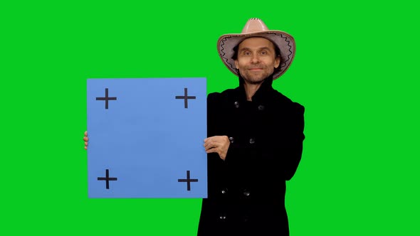 Stylish Man Wearing Black Coat and Cowboy Hat Standing with Blue Blank Board, Chroma Key