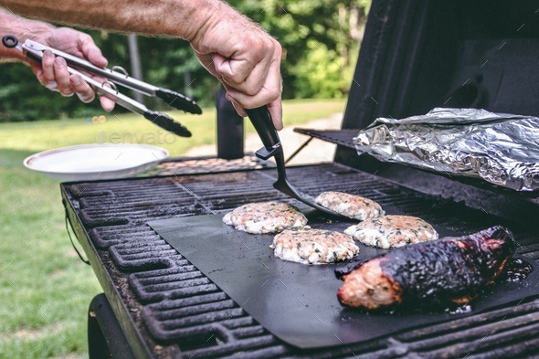 A man grills a pork tenderloin using a grill mat. veggies on the top rack. Salmon burger patties.