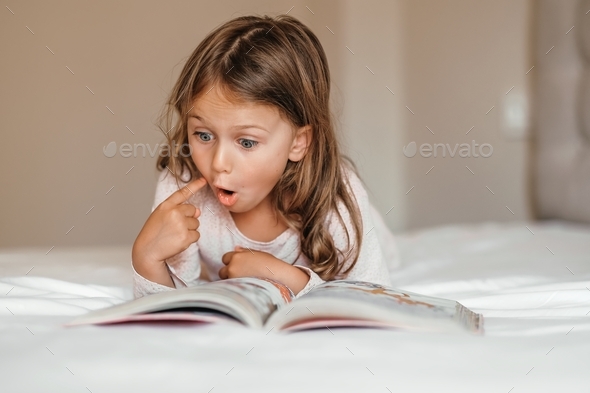 Surprised child girl read book on bed in evening before go to sleep