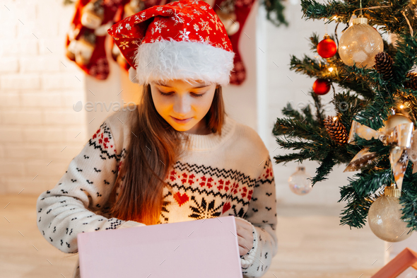 Girl in a red Santa hat opens a gift at home in the living room - Stock Photo - Images