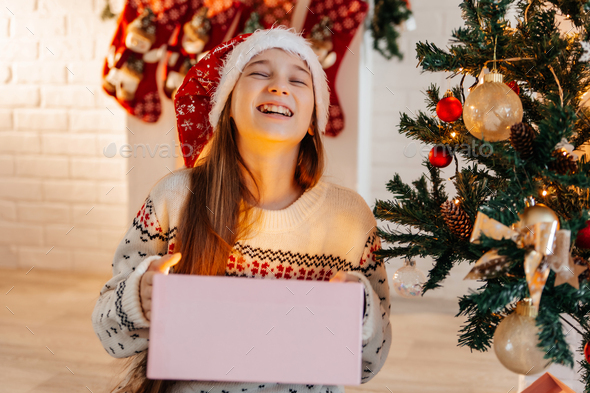 Girl in a red Santa hat opens a gift at home in the living room - Stock Photo - Images