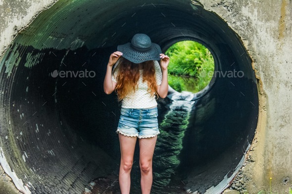 Young woman fashionista tipping hat standing in circular tunnel with streaming water keeping cool