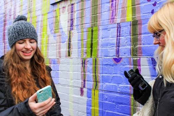Two young women in winter attire by colorful wall using social media on cell phones taking selfies
