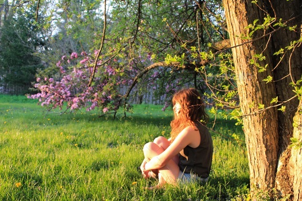 Young woman meditating on moment in nature by floral tree in tall green grass thinking & reflecting