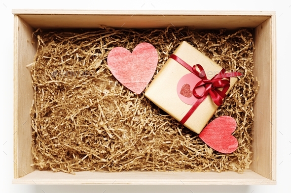 Wooden box with present wrapped in brown craft paper with red ribbon, filled with paper filler