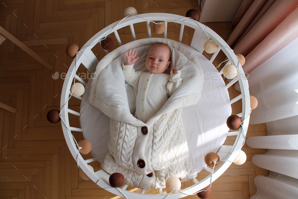 Cute baby in a beige bodysuit lies on back in a round crib with a flashlights.