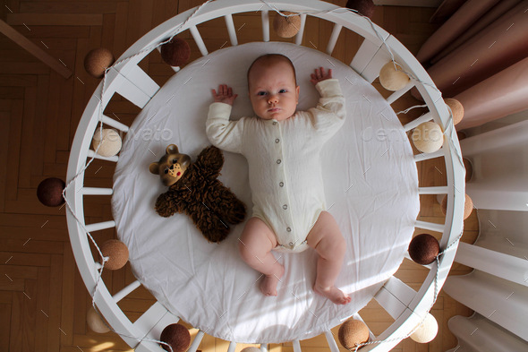 Baby in a beige bodysuit lies on his back on white bed linen in a white round crib with a bear toy