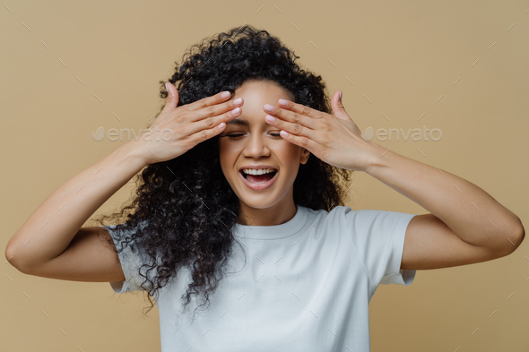 Horizontal shot of happy dark skinned woman covers eyes and smiles, has fun and hides face