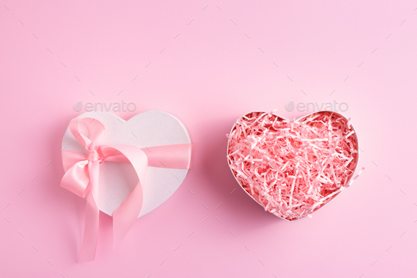 Festive heart shaped present box with pink shred paper on pastel pink background.