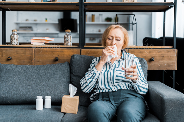 sick senior woman with runny nose sitting on couch with tissue box and holding glass of water in