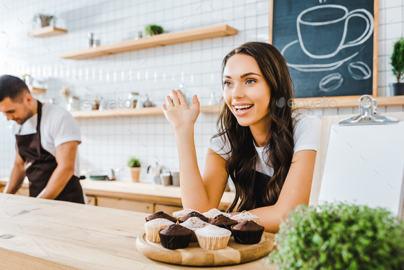 selective focus of attractive brunette waitress standing behind bar counter with cupcakes and waving