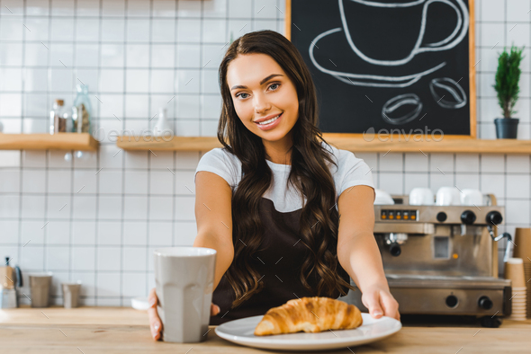 attractive brunette cashier standing behind bar counter with cup, plate and croissant in coffee