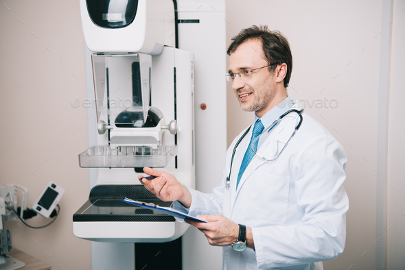 smiling radiographer standing near x-ray machine and holding clipboard ...