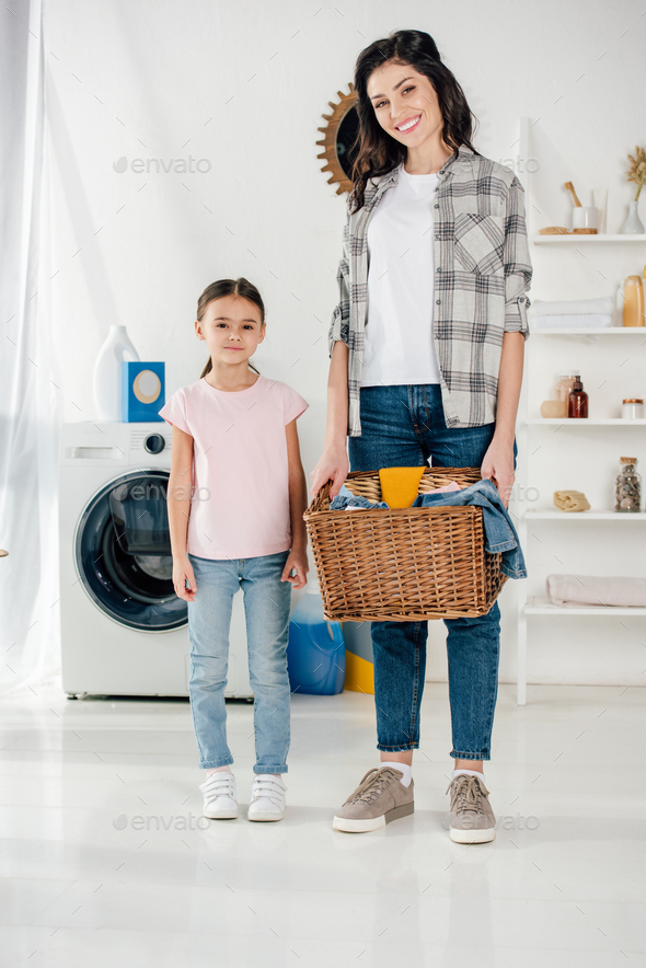 daughter in pink t-shirt standing near smiling mother in grey shirt with basket and in laundry room