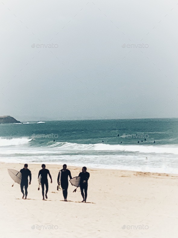 Four surfers walking with their boards against a golden sandy beach with rough surf on the beach