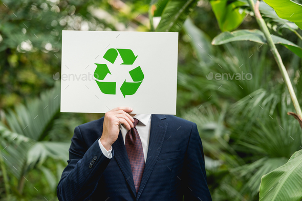 businessman holding white card with green recycling sign in front of face in greenhouse