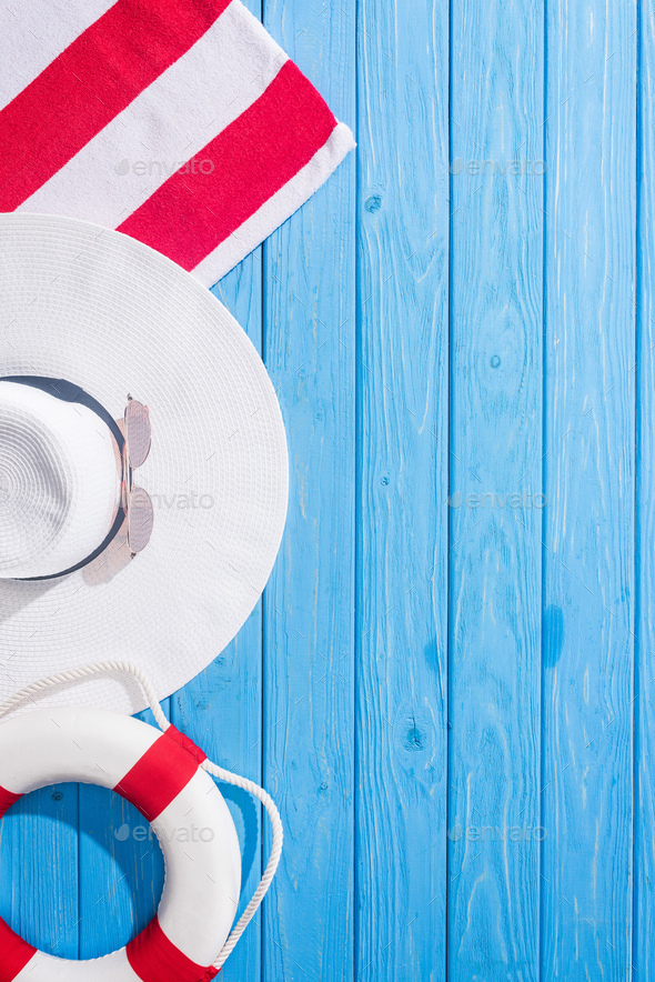 top view of striped towel, white sunglasses, lifebuoy, floppy hat on blue wooden background with