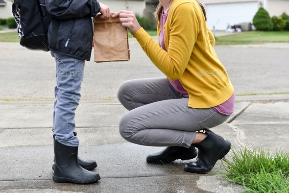 Mother kneeling down in a driveway and handing her child a brown paper bag lunch for school