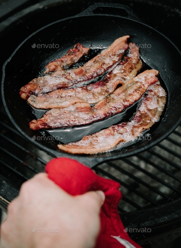 Cooking bacon in a cast iron skillet at a campsite.  - Stock Photo - Images