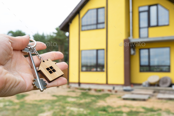 A hand with the keys to a new house on the background of an unfinished cottage