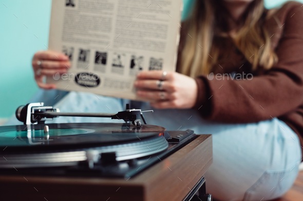 Young woman sitting in front of retro record player holding a record sleeve and listening to music