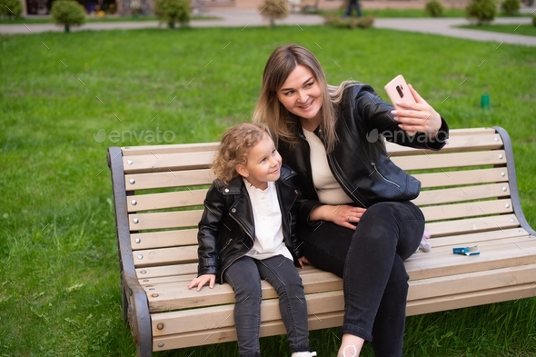 A cute girl in a black jacket is sitting on a bench with her mom laughing and taking selfies