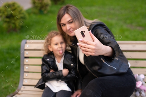 A charming girl in a black jacket sits on a bench with her mom and looks at the phone and shows her