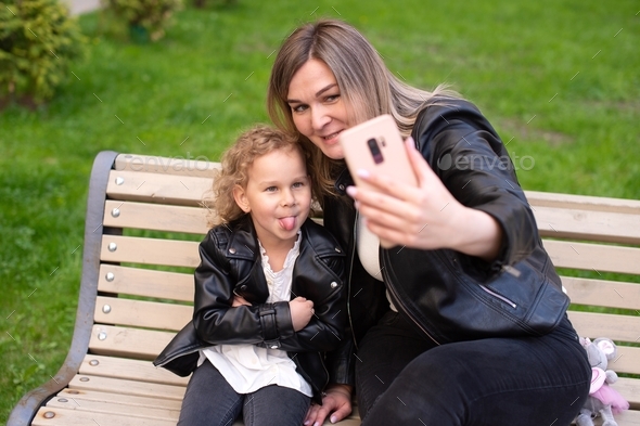 A pretty girl is sitting on a bench with her mom, laughing, taking a selfie and showing her tongue