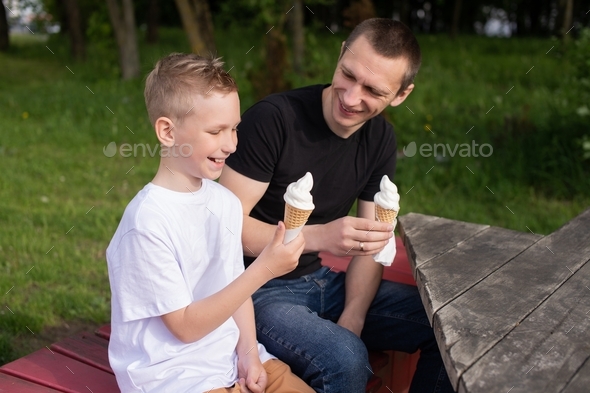 A cute boy in a white T-shirt is sitting with his dad in the park and holding ice cream.