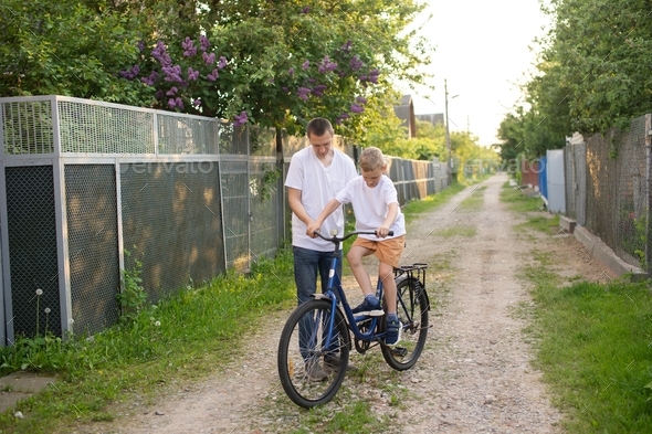 A cute boy in a white T-shirt learns to ride a bike with his dad and laughs. Dad teaches his son.