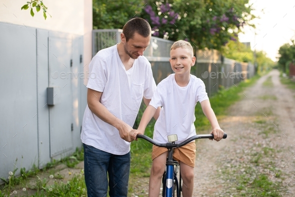A cute boy in a white T-shirt learns to ride a bike with his dad and laughs.