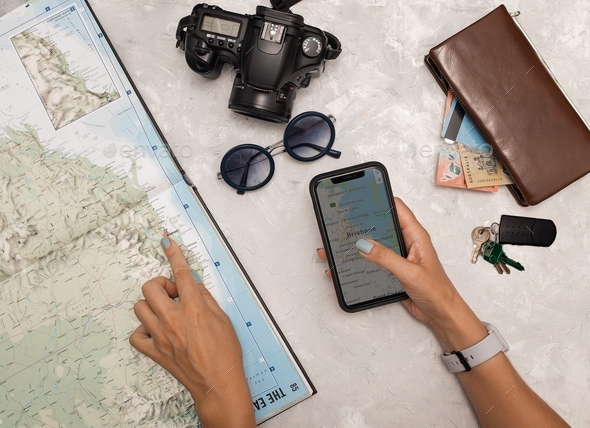 Travel flatlay. Woman’s hands on maps in atlas and mobile phone, car key, wallet, camera, sunglasses