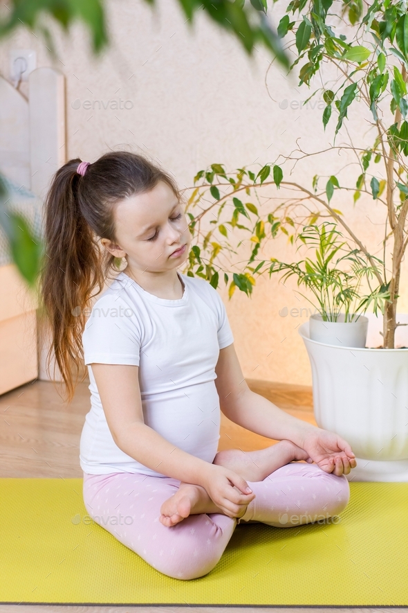 Cute little girl sitting on fitness mat in a lotus position while doing yoga  and meditation at home. Stock Photo by amihay982
