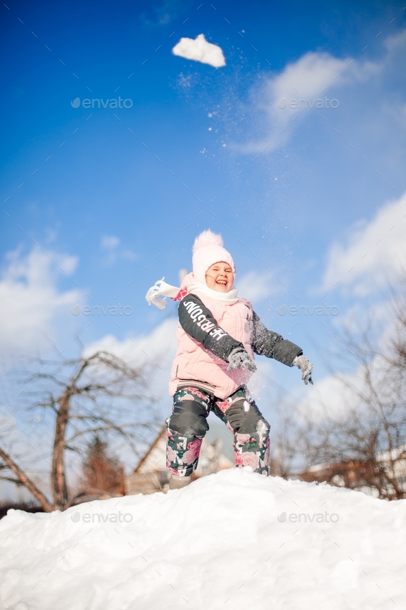 Child throws snowballs in winter park. Little girl in pink winter suit plays and rides down snow