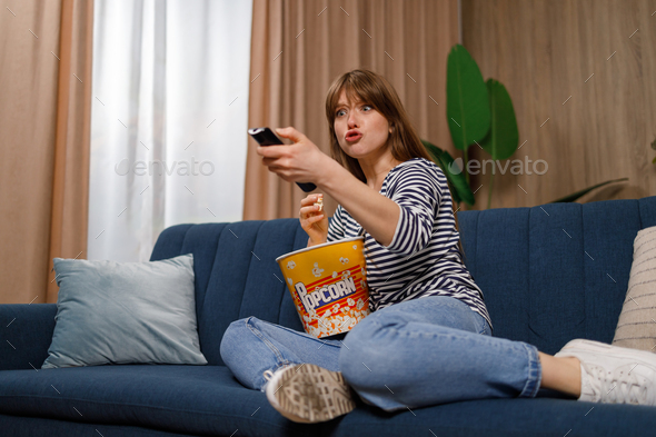 Scared woman with a bucket of popcorn switches channels while watching TV indoor
