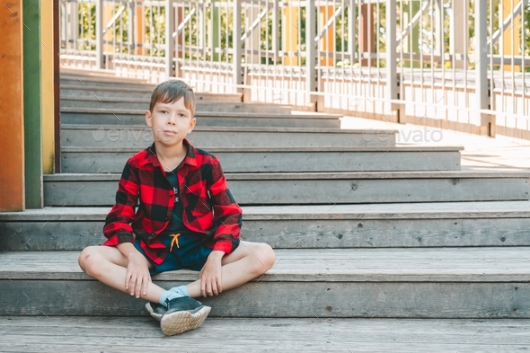 sad boy in a red plaid shirt sits on the stairs in the street. Change in the street, at school.